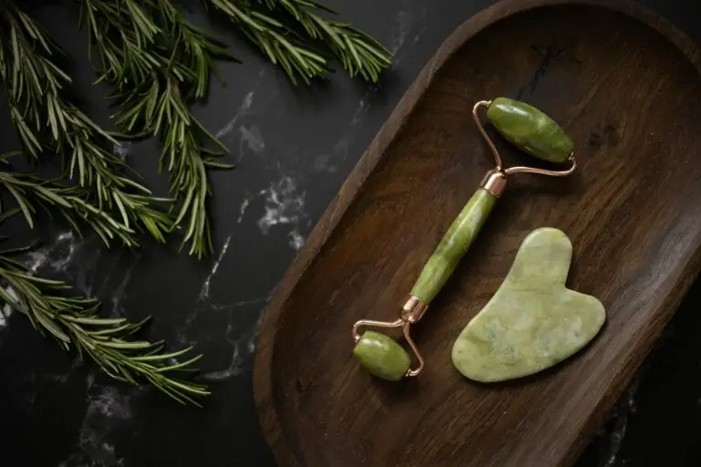 Jade roller and gua sha on dark wood tray with rosemary branches for spa and relaxation.