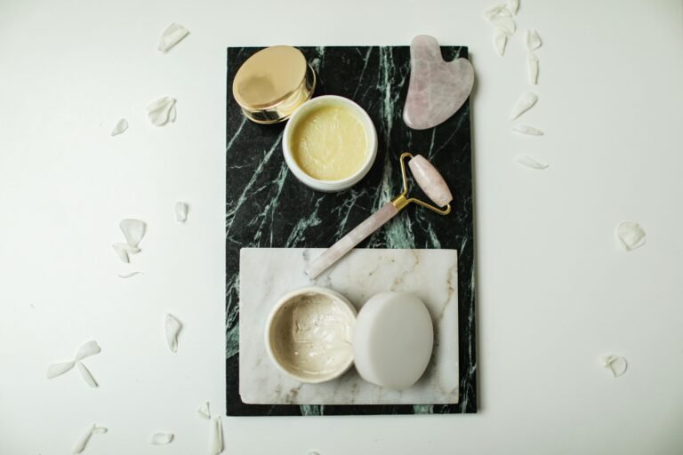 Flatlay of luxurious skincare tools and creams on marble slabs, surrounded by flower petals.