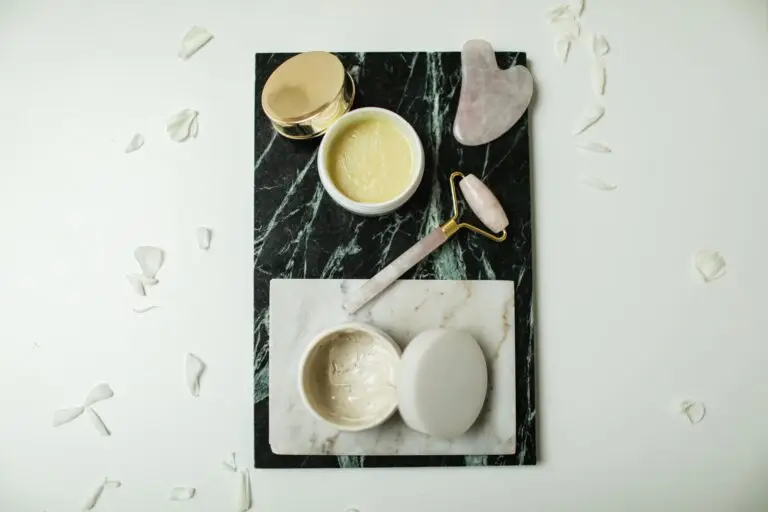 Flatlay of luxurious skincare tools and creams on marble slabs, surrounded by flower petals.